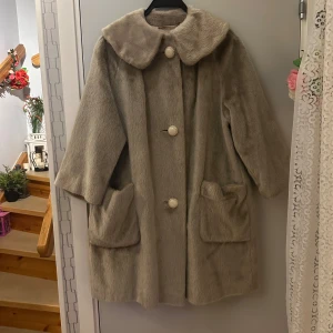 Beige pälsjacka med stora knappar - Säljer en elegant beige pälsjacka med stora dekorativa knappar och bred krage. Jackan har en klassisk design med två stora fickor framtill och trekvartsärmar. Perfekt för kyliga dagar när du vill känna dig både varm och stilfull.                     Passar storlek M/L