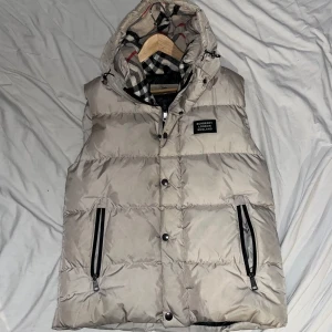 Beige dunväst från Burberry - Snygg beige dunväst från Burberry med klassiskt rutigt foder i huvan. Västen har dragkedja och knappar framtill samt praktiska fickor med dragkedja. Perfekt för kyliga dagar. Priset är diskuterbart.