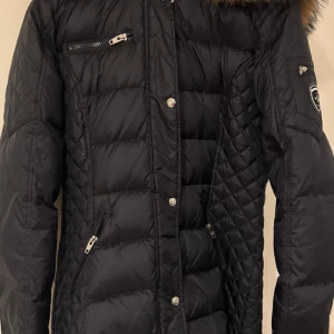 Svart dunjacka med pälsdetaljer ifrån märket rock and blue. - Snygg svart dunjacka från Rockandblue med pälsdetaljer på huvan. Det är äkta päls på luvan.  Jackan har dragkedja och knappar framtill samt flera praktiska fickor. Perfekt för kyliga dagar. Jackan är i nyskick och kostar 2800kr i ursprungs pris. 