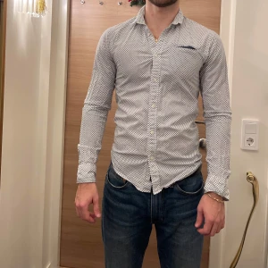 Vit och blå mönstrad skjorta från Scotch & Soda - Snygg vit skjorta med blått mönster från Scotch & Soda. Skjortan har en slim passform och långa ärmar. Perfekt för en stilren look med knappar framtill och en klassisk krage.