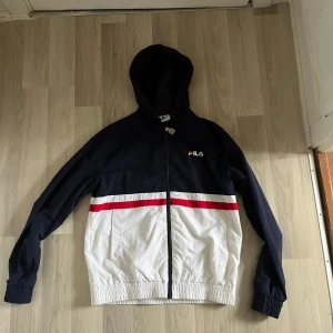Svart och vit vindjacka från FILA - Snygg vindjacka från FILA i svart och vitt med röda detaljer. Jackan har en huva och dragkedja framtill. Perfekt för en sportig look.