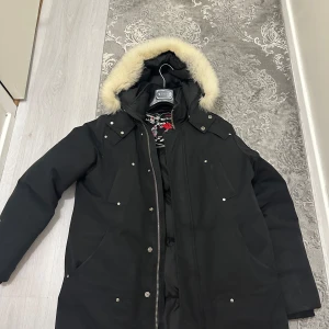 Svart parkajacka med pälsdetalj från Moose Knuckles - Snygg svart parkajacka från Moose Knuckles med avtagbar huva och pälsdetalj. Jackan har dragkedja och knappar framtill samt flera praktiska fickor. Perfekt för kalla dagar.