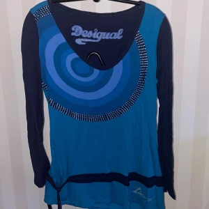 Blå långärmad tröja från Desigual - Snygg blå långärmad tröja från Desigual med ett unikt cirkelmönster i olika blå nyanser och prickiga detaljer. Perfekt för dig som vill sticka ut med stil. Tröjan har en bekväm passform och är gjord i mjukt material.