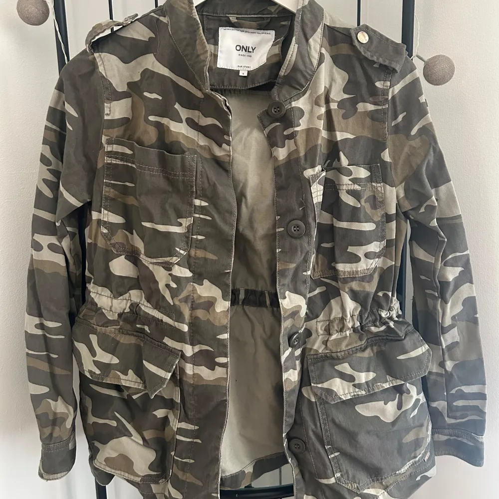 Säljer en cool camouflagejacka från ONLY med knappar och flera fickor. Jackan har en normal passform och är perfekt för en avslappnad stil. Den är långärmad och har en klassisk militärlook.. Takit.