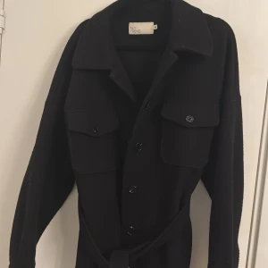 Svart kappa från Nelly - Snygg svart kappa från New Trend med knappar och bälte i midjan. Perfekt för höst och vår med sina långa ärmar och stilrena design. Köpt för 699kr. Passar även S.