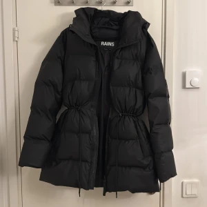 Svart pufferjacka från Rains - Snygg svart pufferjacka från Rains med dragkedja och justerbar midja. Perfekt för kyliga dagar med sin vadderade design och långa ärmar. Jackan har en stilren look och är både praktisk och trendig. Köpt för 3999kr.