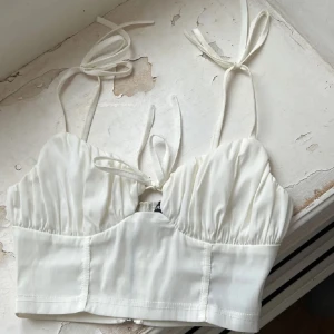 Vit korsett med knytband - Snygg vit korsett med knytband vid axlarna och dragkedja i ryggen. Perfekt för en stilren look. Den har en elegant design med lätt rynkade kupor.