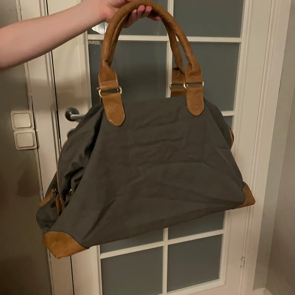 Snygg och rymlig weekendväska i gått tyg med bruna skinnhandtag och detaljer. Väskan har ett randigt innerfoder och en praktisk dragkedja. Perfekt för både vardag och resa.. Laukut & Käsilaukut.