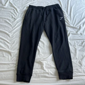 Svarta mjukisbyxor från Gymshark - Säljer ett par svarta mjukisbyxor från Gymshark med elastisk midja och snörning. Byxorna har en avslappnad passform och ribbade muddar vid bensluten. Perfekta för en bekväm dag hemma eller på gymmet.