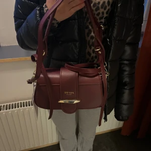 Vinröd axelväska  - Snygg vinröd axelväska  i skinn med gulddetaljer. Väskan har en justerbar axelrem och ett elegant spänne framtill. Perfekt för att ge en lyxig touch till din outfit.