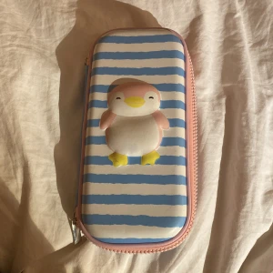 Blå- och vitrandig pennfodral med pingvinmotiv - Sött pennskrin med blå- och vitrandigt mönster och ett gulligt pingvinmotiv på framsidan. Insidan är rosa med flera fack och en dragkedja för extra förvaring. Perfekt för att ha fina pennor i!💕