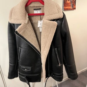 Svart jacka med teddyfoder från Stradivarius - Snygg svart jacka från Stradivarius med beige teddyfoder. Jackan har en asymmetrisk dragkedja och bred krage. Perfekt för kyligare dagar med sin stilrena design och bekväma passform.