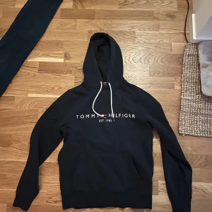 Mörkblå hoodie från Tommy Hilfiger - Säljer en stilren marinblå hoodie från Tommy Hilfiger med vit dragsko och logotyp. Säljer pga för liten för mig. Bra skick och inga synliga defekter man kan hitta. Köpt från boozt 1200kr. Skriv om du har frågor om pris eller annat!