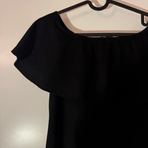 Svart offshoulder klänning från Nly One - Elegant svart offshoulder klänning från Nly One i storlek XS. Klänningen har en stilren design med volangdetaljer vid axlarna och en figurnära passform som ger en smickrande siluett.