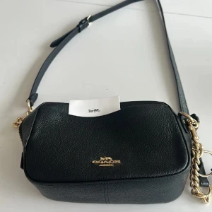 Svart axelväska från Coach - Snygg svart axelväska från Coach i skinn med guldfärgade detaljer. Väskan har en justerbar axelrem och en dragkedja för säker förvaring. Perfekt för att bära dina nödvändigheter med stil.