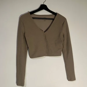 Beige ribbad kofta - Säljer en stilren beige ribbad kofta med v-ringning och knapp framtill. Den är croppad och har långa ärmar, perfekt för lager på lager-stil. Har använts endast 1 gång. 