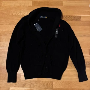 Svart Cardigan från Polo Ralph Lauren - Snygg svart kofta från Polo Ralph Lauren med knappar framtill. Den har en klassisk design med ribbade detaljer och med röd logga, Storlek XL.       Står ej för returer eller postnordslarv