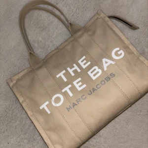 Beige Tote Bag från Marc Jacobs - Snygg beige tote bag från Marc Jacobs med texten 'THE TOTE BAG' tryckt i vitt. Väskan har två handtag och en dragkedja upptill för säker förvaring. Perfekt för att bära med sig allt man behöver under dagen.