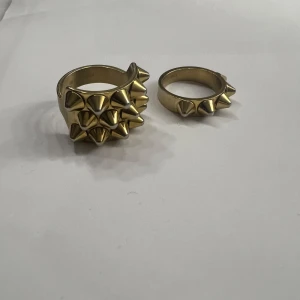 Guldiga ringar med nitar - Två coola guldiga ringar med nitar. Den ena ringen har en bredare design med flera rader av nitar, medan den andra är smalare med en enkel rad. Perfekta för att ge en edgy touch till din stil.