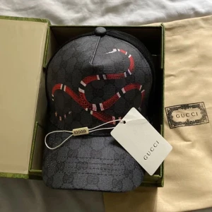 Svart keps med ormmönster från Gucci - Snygg svart keps från Gucci med ett iögonfallande ormmönster i rött och vitt. Kepsen har en klassisk design med böjd skärm och justerbar passform.