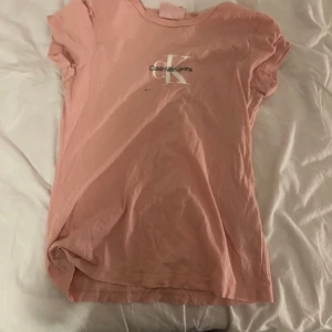 Rosa t-shirt från Calvin Klein - Snygg rosa t-shirt från Calvin Klein med deras ikoniska logga på bröstet. Perfekt för en avslappnad stil. Kortärmad och i mjukt bomullsmaterial.