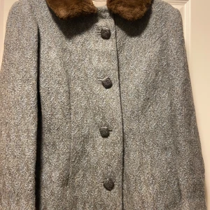 Grå kappa med pälsdetaljer - Elegant grå kappa med bruna pälsdetaljer på kragen. Kappan har dekorativa knappar och är perfekt för kyliga dagar. Den har en klassisk design som passar bra till både vardag och mer formella tillfällen.