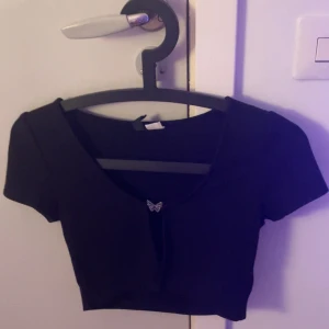 Svart croppad topp med fjärilsdetalj - Snygg svart croppad topp från H&M med en liten fjärilsdetalj framtill. Toppen har korta ärmar och en djupare urringning. Står ej för frakten och kan gärna pröva den och skicka bild ☺️❤️