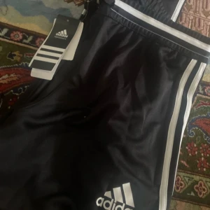 Svarta träningsbyxor från Adidas - Snygga svarta träningsbyxor från Adidas med vita ränder längs sidorna. Byxorna har en elastisk midja och är perfekta för sport och träning. Adidas-loggan är tryckt på framsidan.