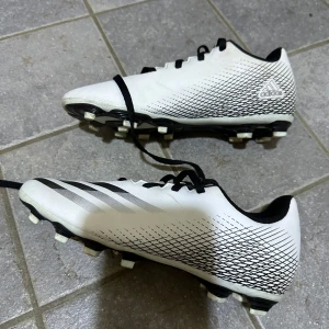 Vita fotbollsskor från Adidas - Säljer ett par vita fotbollsskor från Adidas med svarta detaljer och snörning. Skorna har en mönstrad yta och dobbar för bra grepp på planen. Perfekta för fotbollsträning och matcher.