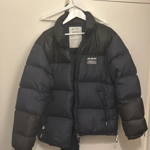 Svart och blå pufferjacka från Axel Arigato - Säljer en snygg svart och blå pufferjacka från Axel Arigato. Jackan har en dragkedja framtill och är långärmad. Perfekt för kyliga dagar.