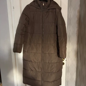 Brun lång dunjacka med huva - Säljer en brun lång dunjacka med huva. Jackan har en dragkedja framtill och långa ärmar. Perfekt för kyliga dagar. I bra skick, har använts ofta men känns fortfarande ny!