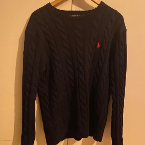 Mörkblå tröja från Polo Ralph Lauren - Säljer en mörkblå kabelstickad tröja från Polo Ralph Lauren. Tröjan har en klassisk design med rund hals och det ikoniska röda logotypbroderiet på bröstet. Perfekt för en elegant och avslappnad look.