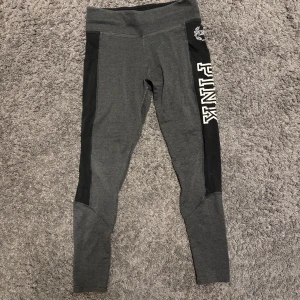 Victoria’s secret PINK - Snygga grå leggings från PINK med svarta paneler längs sidorna och logotypen tryckt i vitt. Perfekta för träning eller en avslappnad dag. De har en hög midja för extra komfort.