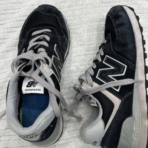 Svarta sneakers från New Balance - Snygga svarta sneakers från New Balance med grå detaljer och snörning. Skorna har en klassisk design med det ikoniska 'N' på sidan. Perfekta för en avslappnad stil.