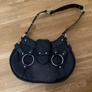 Svart snygg axelväska med detaljer.  - Snygg svart axelväska med silverfärgade ringdetaljer och nitar. Väskan har en justerbar axelrem och stängs med dragkedja. Perfekt för att ge en edgy touch till din outfit och har använt få tal gånger. 😁❤️