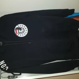 Svart hoodie från Dsquared2 - Säljer en svart hoodie från Dsquared2 med ett stort tryck på ryggen och ett runt emblem på framsidan. Hoodien har långa ärmar och en justerbar huva med dragsko. Perfekt för en cool och avslappnad stil.