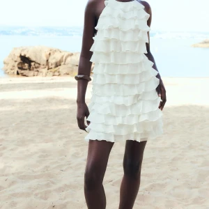 SÖKER  - Säljer en söt vit klänning med flera lager av volanger. Klänningen är ärmlös och har en knytning i ryggen, vilket ger en luftig och somrig känsla. Perfekt för en dag på stranden eller en sommarfest.