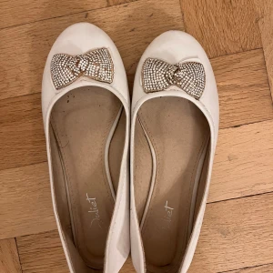 Vita ballerinaskor med glittrig rosett - Eleganta vita ballerinaskor prydda med en glittrig rosett på tån. Perfekta för en stilfull look.