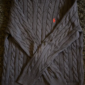 kabelstickad tröja från Ralph Lauren - Säljer en stilren marinblå kabelstickad tröja från Ralph Lauren med det klassiska röda logotypen på bröstet. Tröjan har långa ärmar och ribbade muddar vid ärmslut och nederkant. Perfekt för kyliga dagar! Köpt för 2395 från Zalando.