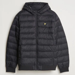 Svart pufferjacka från Lyle & Scott - Snygg svart pufferjacka från Lyle & Scott med dragkedja och huva. Jackan har ett diskret gult logomärke på bröstet och ribbade muddar vid ärmslut och nederkant. Perfekt för kyliga dagar.