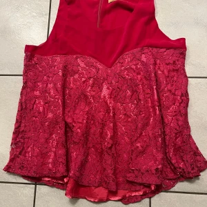 Röd/rosa spets topp - Snygg röd klänning med spetsdetaljer och en elegant design. Klänningen har en ärmlös stil och en dragkedja i ryggen för enkel påklädning. Perfekt för festliga tillfällen.