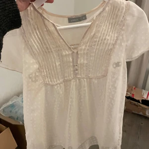 Beige blus från Lindex - Säljer en söt beige blus från Lindex i storlek 36. Blusen har korta ärmar och ett diskret prickigt mönster. Den har en v-ringad design med knappar framtill och fina veckade detaljer på bröstet.
