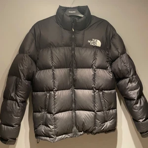 Svart dunjacka från The North Face - Säljer en svart dunjacka från The North Face med dragkedja och hög krage. Jackan har en pufferdesign och är perfekt för kalla dagar. Logotypen är tryckt på bröstet och ryggen. Passar både för stad och natur.