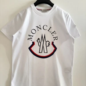 Vit t-shirt från Moncler - Snygg vit t-shirt från Moncler med tryck på framsidan och ett broderat märke på ärmen till ett billigare pris. Perfekt för en stilren och avslappnad look. Storleken är M och kan även passa någon med ett mindre L. Skickas inom 24 timmar.