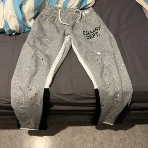 Grå mjukisbyxor från Gallery Dept. - Säljer ett par grå mjukisbyxor från Gallery Dept. med vita och svarta detaljer. Byxorna har en elastisk midja med snörning och är tillverkade i 100% bomull. Perfekta för en avslappnad stil.