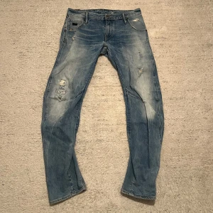 G-Star Jeans - Säljer ett par blå jeans från G-Star med slitna detaljer och en klassisk femficksdesign. Jeansen har en knappgylf och är i en straight fit. Perfekta för en avslappnad stil.