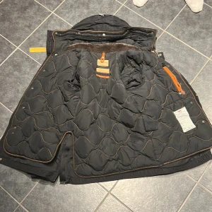 Svart parajumber jacka - Snygg svart PJ jacka med quilted insida och flera praktiska fickor. Jackan har en huva och detaljer i orange och gul. Perfekt för kyligare dagar.