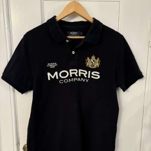 Svart pikétröja från Morris - Snygg svart pikétröja från Morris med broderad logga och text på bröstet. Tröjan har en klassisk krage med knappar och korta ärmar. Perfekt för en stilren look.