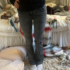 Grå jeans med slitningar - Säljer ett par grå jeans med slitningar på knäna. De har en bootcut-stil och är högmidjade med knappar på bakfickorna. Perfekta för en avslappnad look.
