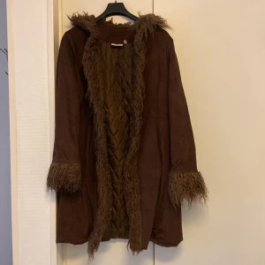 Brun mocka kappa med pälsdetaljer - Snygg brun mocka kappa med quiltat foder och fluffiga pälsdetaljer på huva, ärmslut och framkant. Perfekt för kyliga dagar när du vill hålla stilen. Kappan har en lång och elegant passform. I storlek 40 men passar  mig som har 38. 